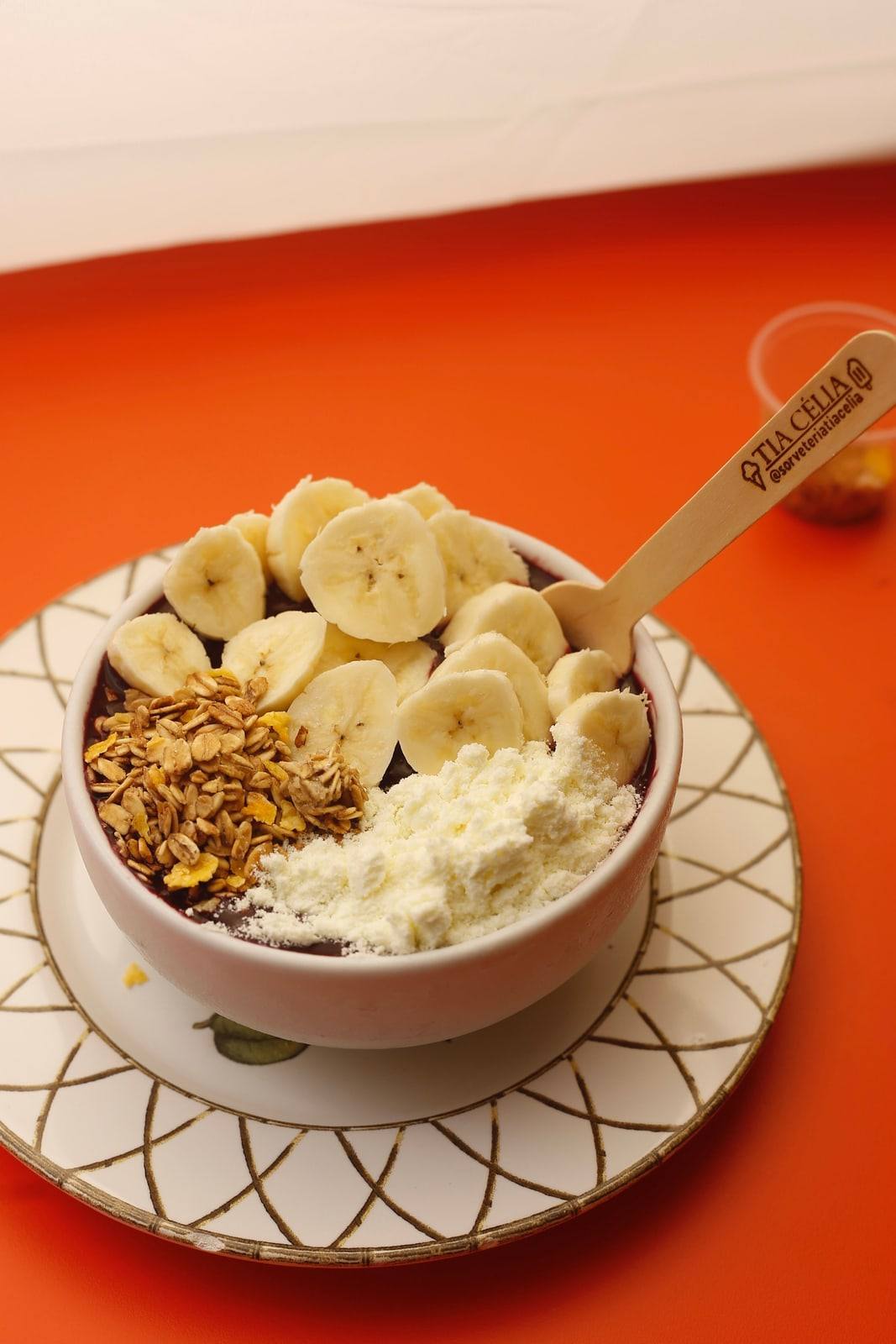 Tigela de açaí com banana, granola e leite em pó, com colherzinha de madeira da Tia Célia, sobre prato decorado em fundo laranja.