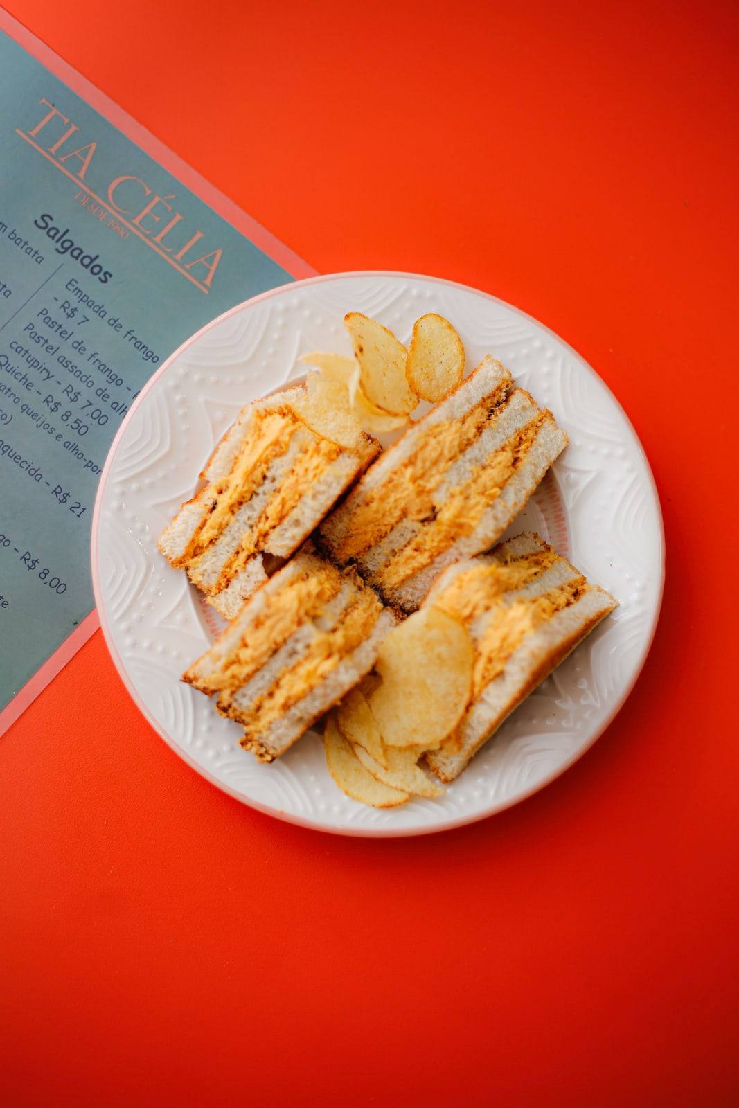 Sanduíche de galinha cortado em triângulos com batata chips, servido em prato branco sobre mesa laranja.