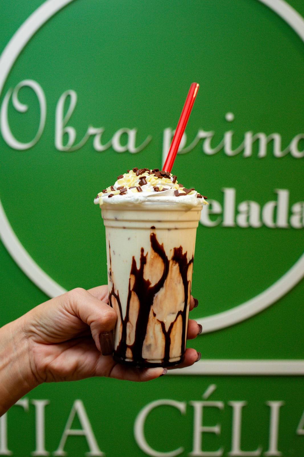 Milk-shake cremoso com chantilly e calda de chocolate escorrendo pelo copo, em frente à parede verde da loja.
