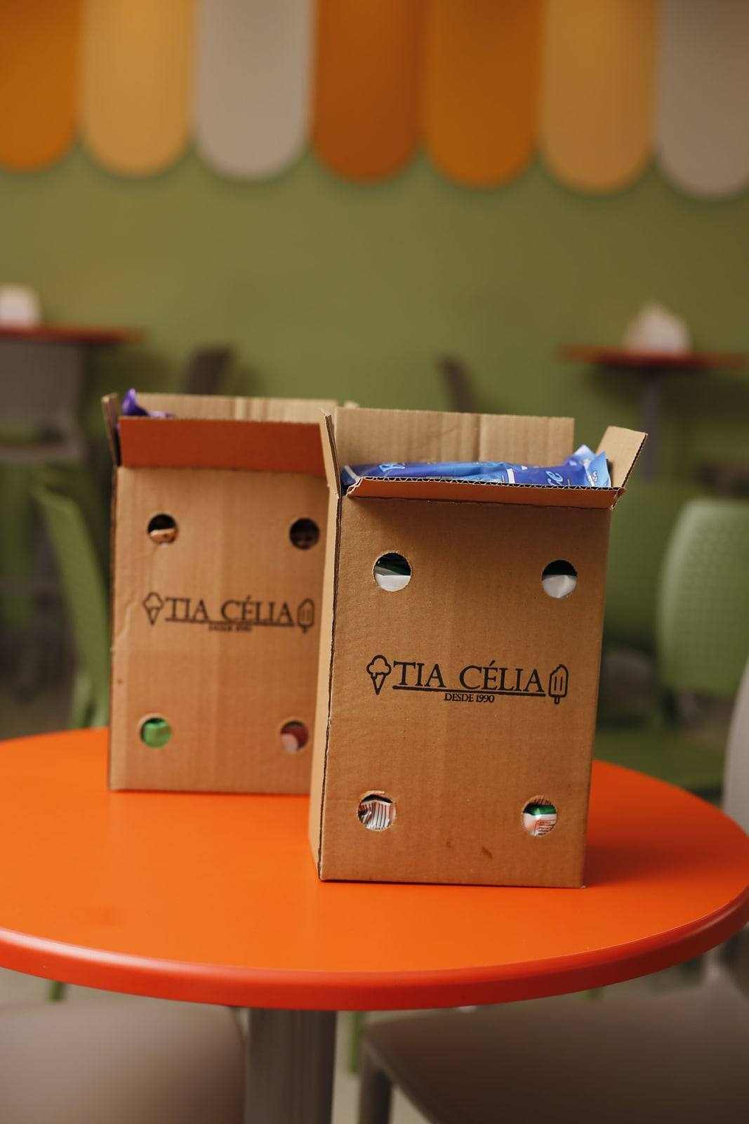 Caixas de papelão da Tia Célia cheias de picolés sobre uma mesa laranja no salão.